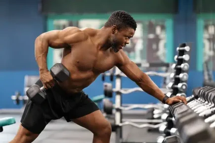 Motivated african american shirtless bodybuilder training arms and back muscles with dumbbells at modern gym, empty space.